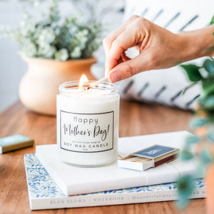 Happy Mother's Day candle being lit by hand holding match sitting on journals surrounded by house plants in brightly lit home on wood grain table 