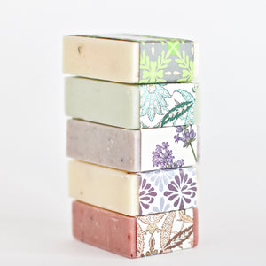 Handmade soap 5 bars stacked on top of each other .  Essential oils soaps wrapped in beautiful letterpress gold flocked artisan papers including  rosemary lemon mint soap warped in green paper, tea tree lime almond soap wrapped in teal paper, oatmeal lavender sage soap wrapped in lavender print paper, lavender lemongrass soap wrapped in purple paper and hibiscus rose geranium soap wrapped in peach and pink paper.