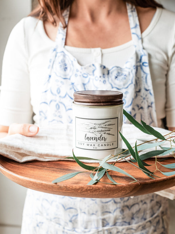 Lavender essential oil candle on an olive wood tray with a botanical sprig of eucalyptus held by a woman wearing a linen apron