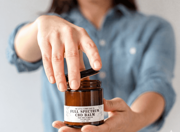 Woman opening a 4oz jar of CBD Cream topical balm for pain reilef CBD/THC balm pain cream rub with black lid in amber glass jar ith 840 mg CBD and 12.6 mg CBC by the little flower soap co