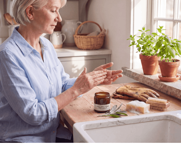 Woman using topical CBD Cream on arthritis hands after gardening CBD rub for pain on a kitchen counter with gardening gloves, soap and a nail brush in a sunny window where basil is growing.