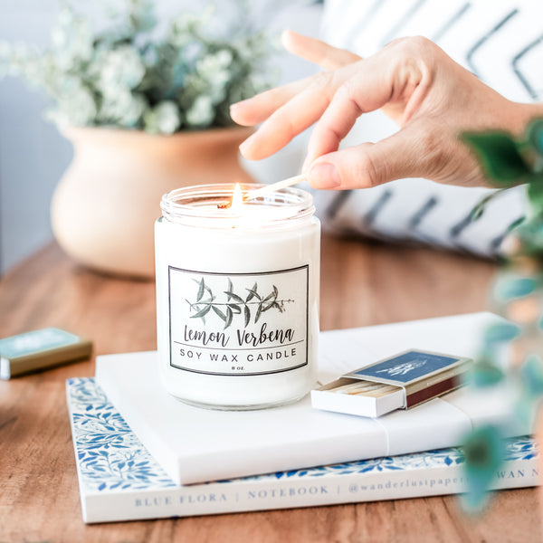 Lemon Verbena soy wax candle white with minimalist black botanical line drawing label and clean fonts for modern farmhouse decor being lit by a match settin on a stack of journals surrounded by plants in a bright light airy space