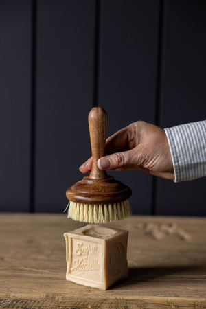 Hand holding the luxe b. high end wooden dish washing brush over a block of soap on a wooden surface with a dark background