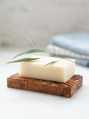 A bar of handmade soap on a gorgeous olive wood hand-carved soap dish a sprig of olive on top and some linen hand towels behind in a brightly lit bathroom