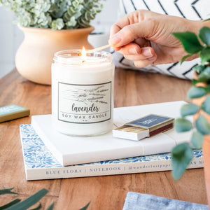 A lavender essential oil soy candle being lit using botanical decorative matches surrounded by sunlight and plants sitting on 2 beautiful journals