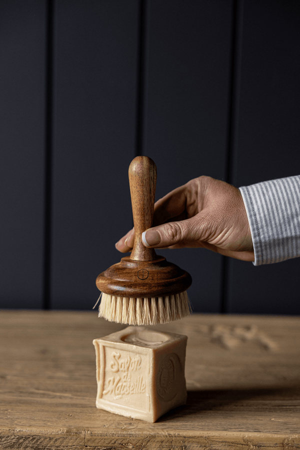 Hand holding the luxe b. high end wooden dish washing brush over a block of soap on a wooden surface with a dark background