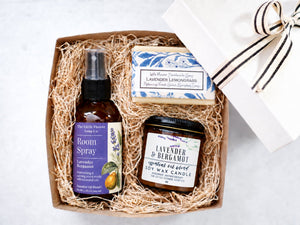 A curated gift box set from The Little Flower Soap Co. containing lavender room spray, a lavender & bergamot soy wax candle, and a lavender lanconglass soap bar, arranged in shredded paper inside a brown kraft box next to a white gift box with a black and white striped ribbon.