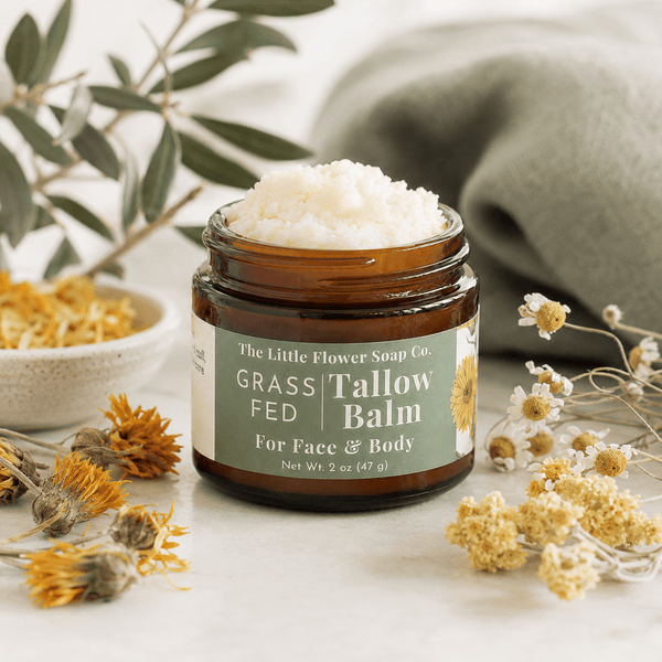Jar of 'Grass Fed Tallow Balm' from The Little Flower Soap Co. on a neutral background with flowers and plants.